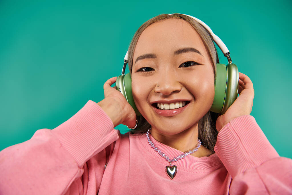 Kablosuz kulaklıklı neşeli ve genç Asyalı kadın turkuaz arka planda müzik dinliyor. - Fotoğraf, Görsel