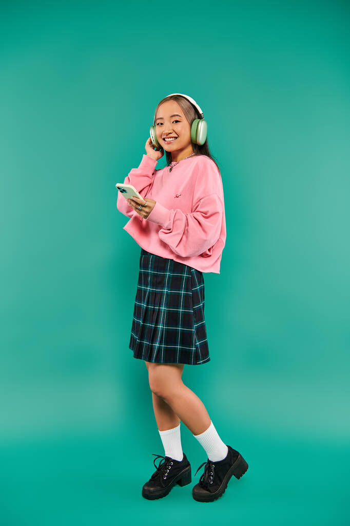 vrolijk aziatisch meisje in draadloze hoofdtelefoon luisteren muziek en met behulp van smartphone op turquoise - Foto, afbeelding