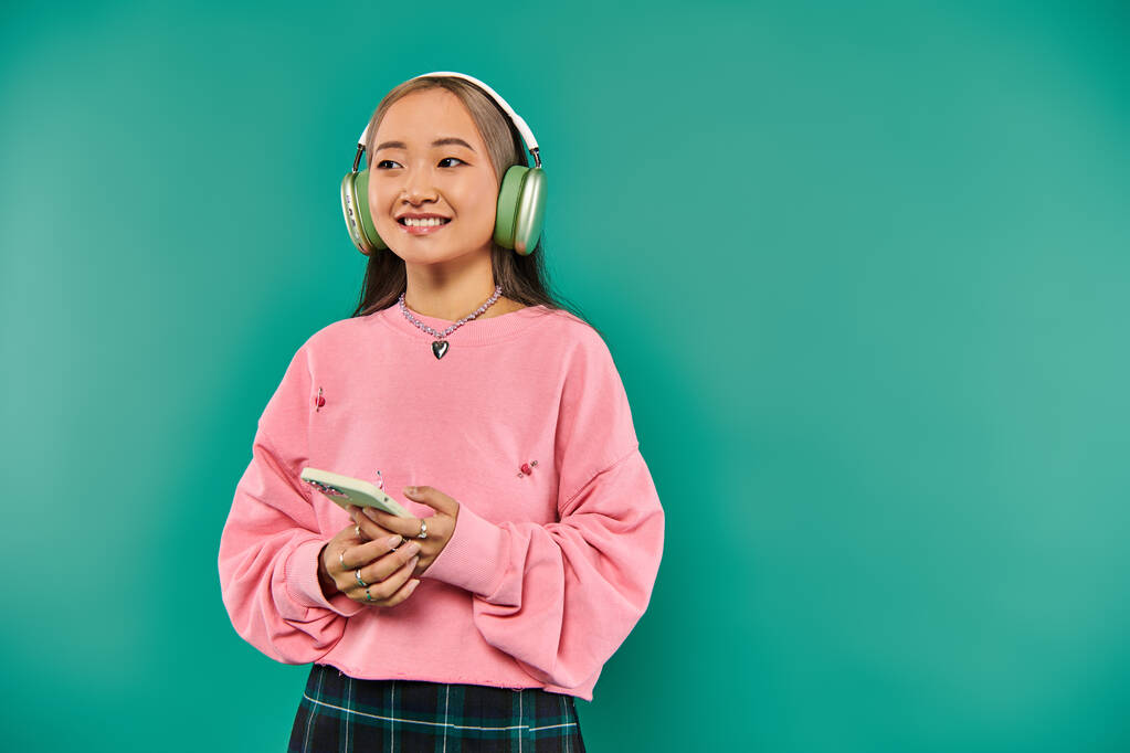 весела азіатка в бездротових навушниках слухає музику і використовує смартфон на бірюзовому фоні - Фото, зображення