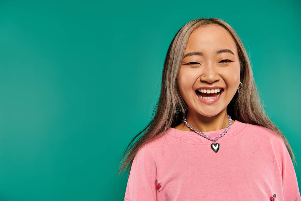 портрет веселой и молодой азиатской девушки в розовой толстовке, позирующей на бирюзовом фоне - Фото, изображение