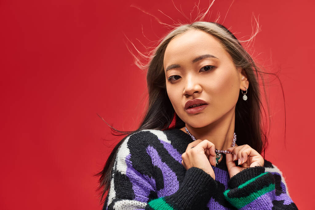 alluring young asian woman in vibrant sweater with animal print touching necklace on red backdrop - Photo, Image