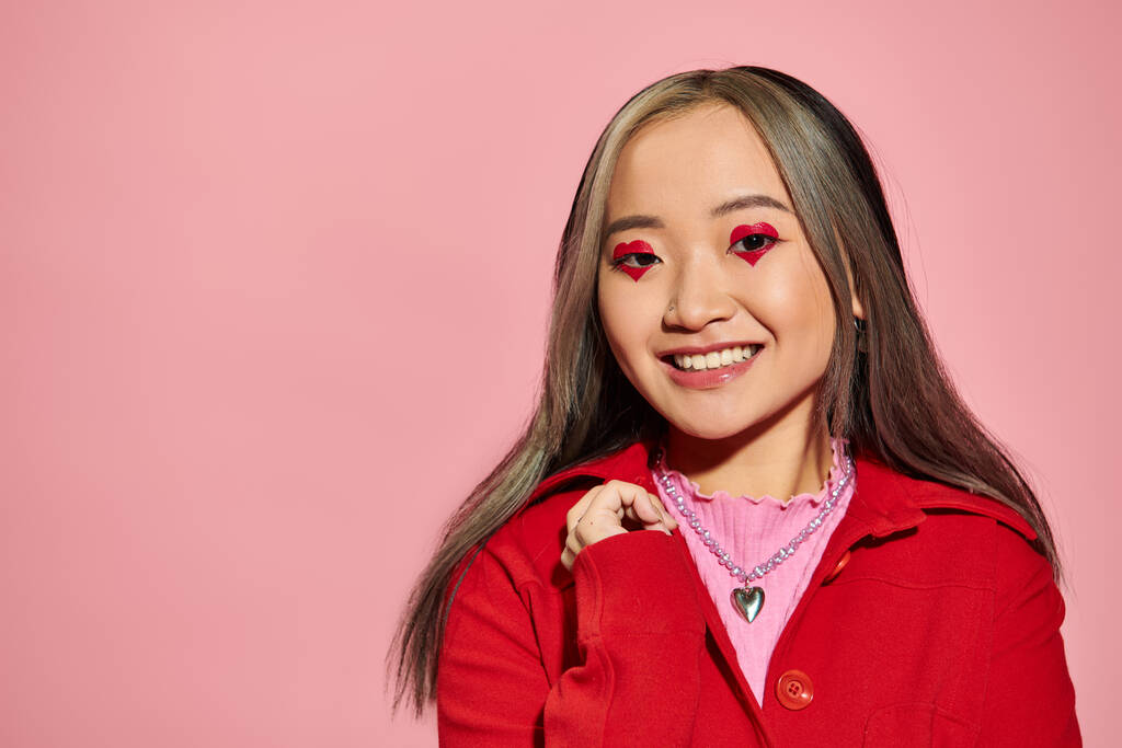 Valentines day concept, cheerful asian woman with heart shaped eye makeup smiling on pink backdrop - Photo, Image