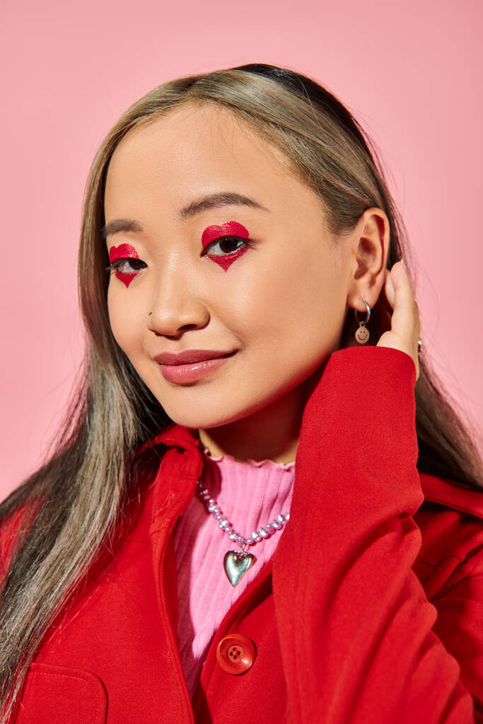 Valentine dia, positivo asiático jovem mulher com coração olho maquiagem posando no vermelho jaqueta no rosa - Foto, Imagem