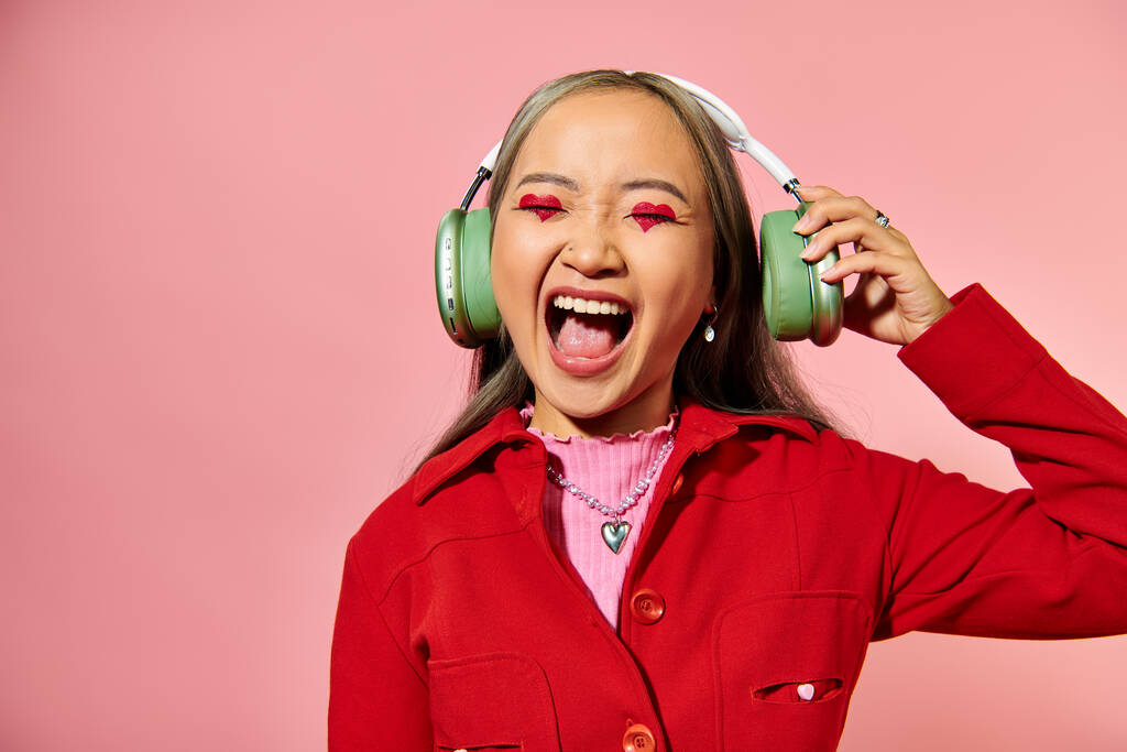 ワイヤレスヘッドフォンで音楽を聞き,ピンクの背景で叫ぶ興奮した若いアジアの女性 - 写真・画像