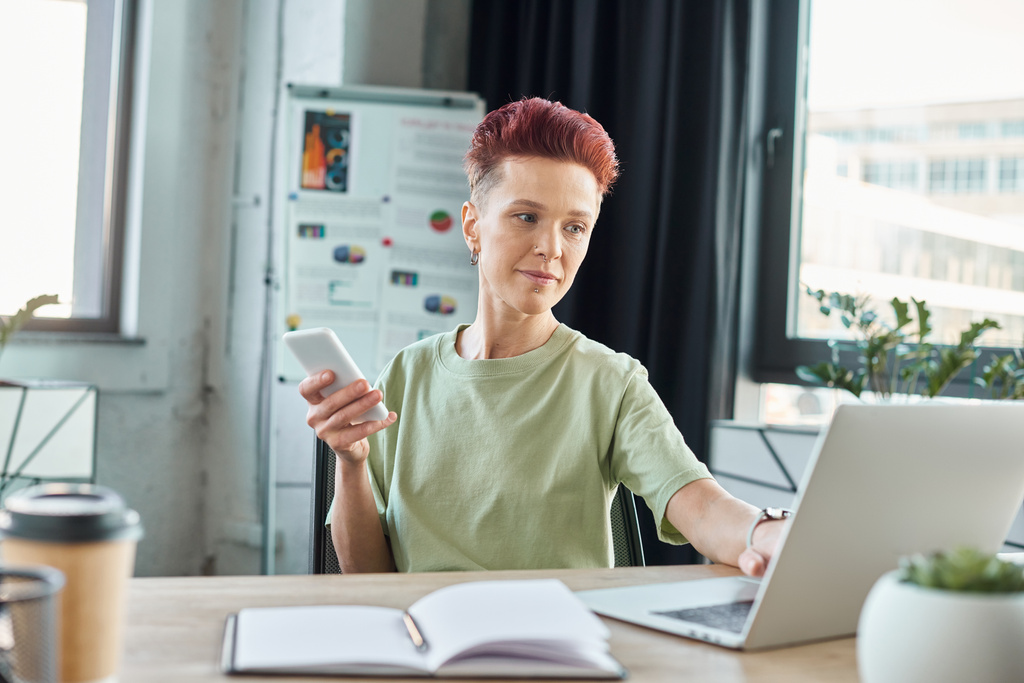 stijlvol bigender individu met smartphone werkt op laptop in de buurt van notebook in modern kantoor - Foto, afbeelding