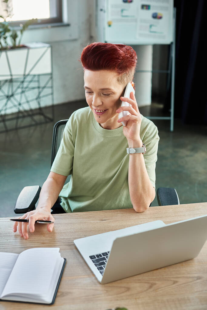 μη δυαδικό πρόσωπο επιχείρησης που μιλά στο κινητό τηλέφωνο κοντά στο lap-top στο χώρο εργασίας στο σύγχρονο γραφείο - Φωτογραφία, εικόνα