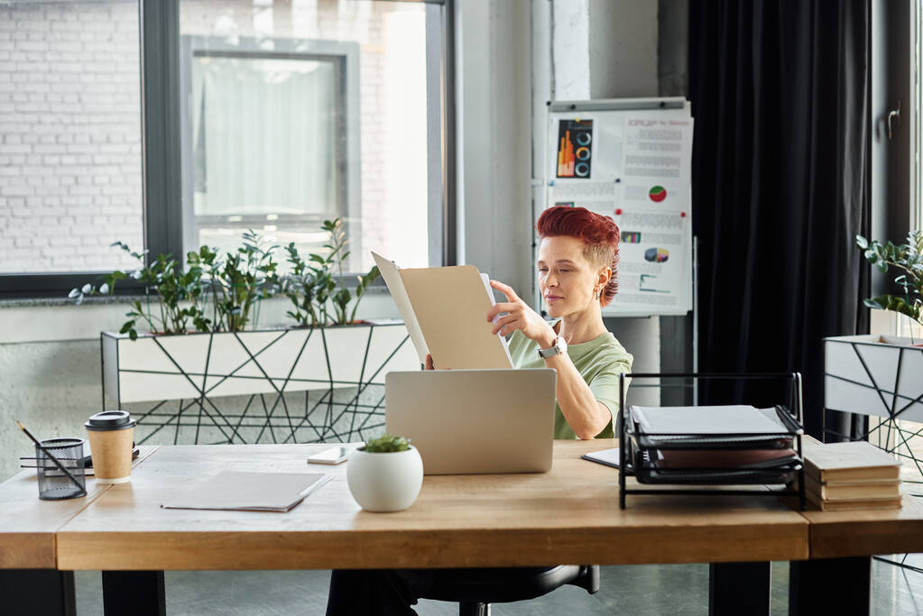 stijlvolle bigender manager werken met documenten in de buurt van laptop en koffie te gaan in papieren beker op kantoor - Foto, afbeelding