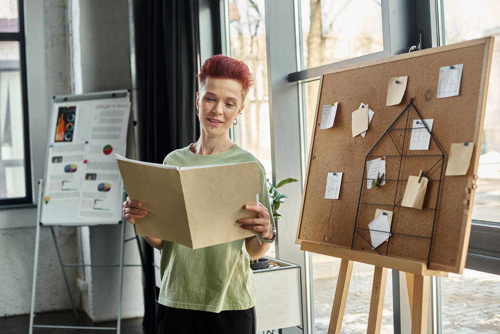 улыбающийся педик смотрит на папку с документами рядом с пробковыми досками с бумажными записками в офисе - Фото, изображение
