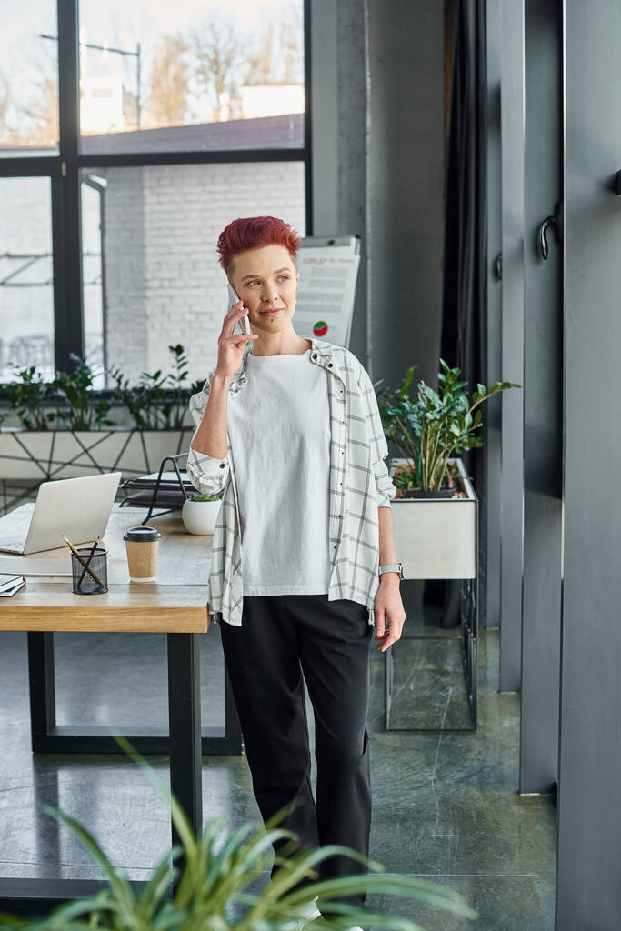 Modern ofiste gündelik kıyafetlerle şık biseksüel menajer ayakta duruyor ve akıllı telefonla konuşuyor. - Fotoğraf, Görsel