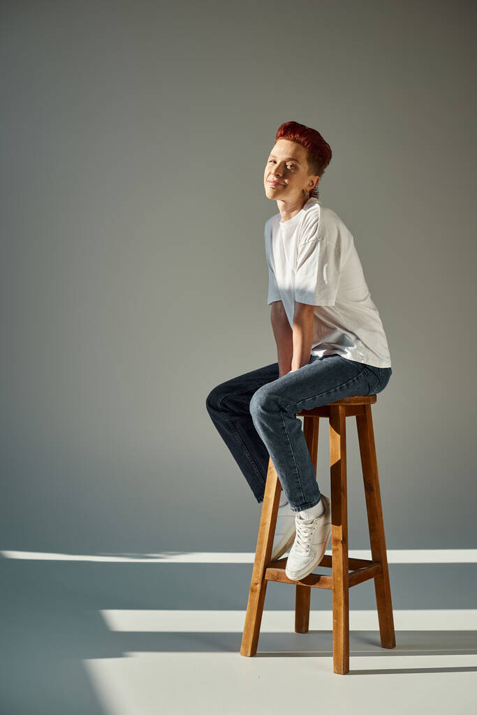 happy non-binary person in white t-shirt and jeans sitting on stool in sunlight on grey, full length - Photo, Image