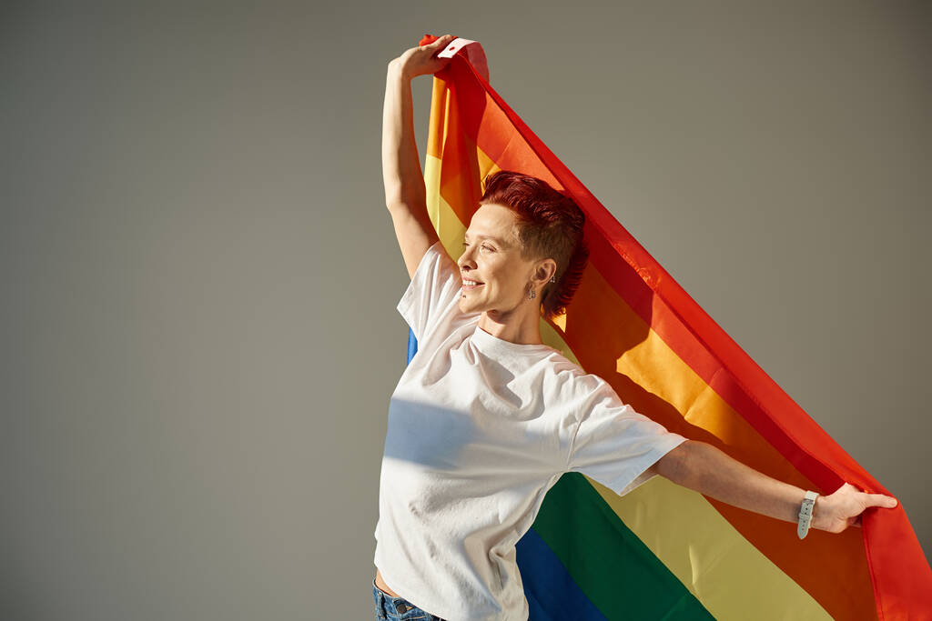 happy and unique queer person in white t-shirt posing with rainbow colors LGBT flag on grey backdrop - Photo, Image