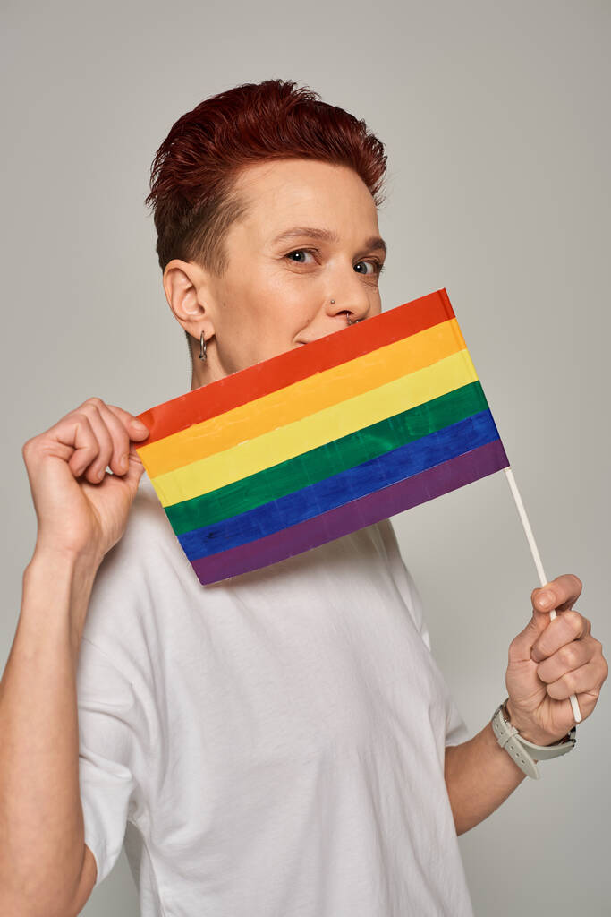 руда квір-модель в білій футболці позує з маленькою ЛГБТ-квартирою біля обличчя, дивлячись на камеру на сірому - Фото, зображення