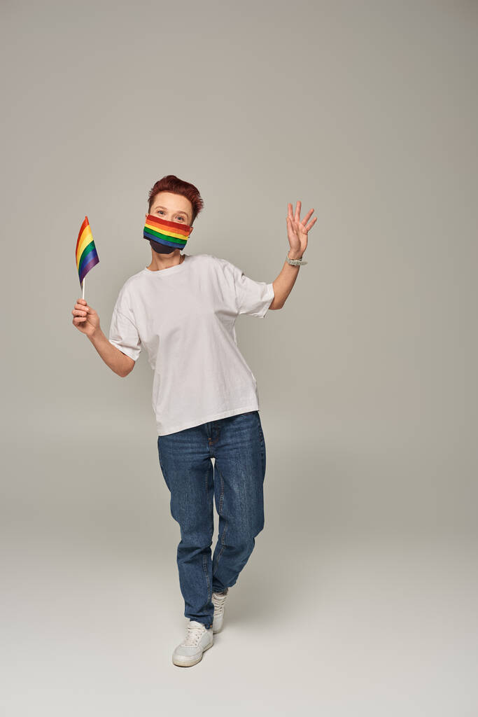full length of redhead queer person in rainbow colors medical mask with small LGBT flag on grey - Photo, Image