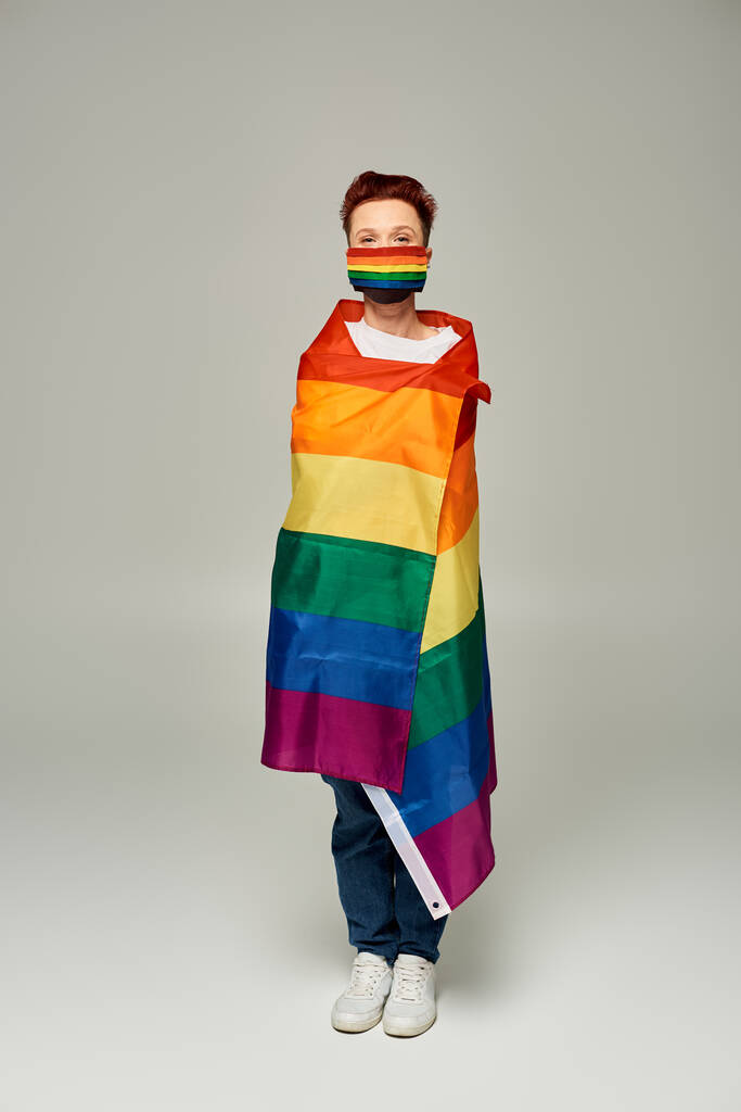 longitud completa de pelirroja queer persona en arco iris colores máscara médica posando en la bandera LGBT en gris - Foto, Imagen