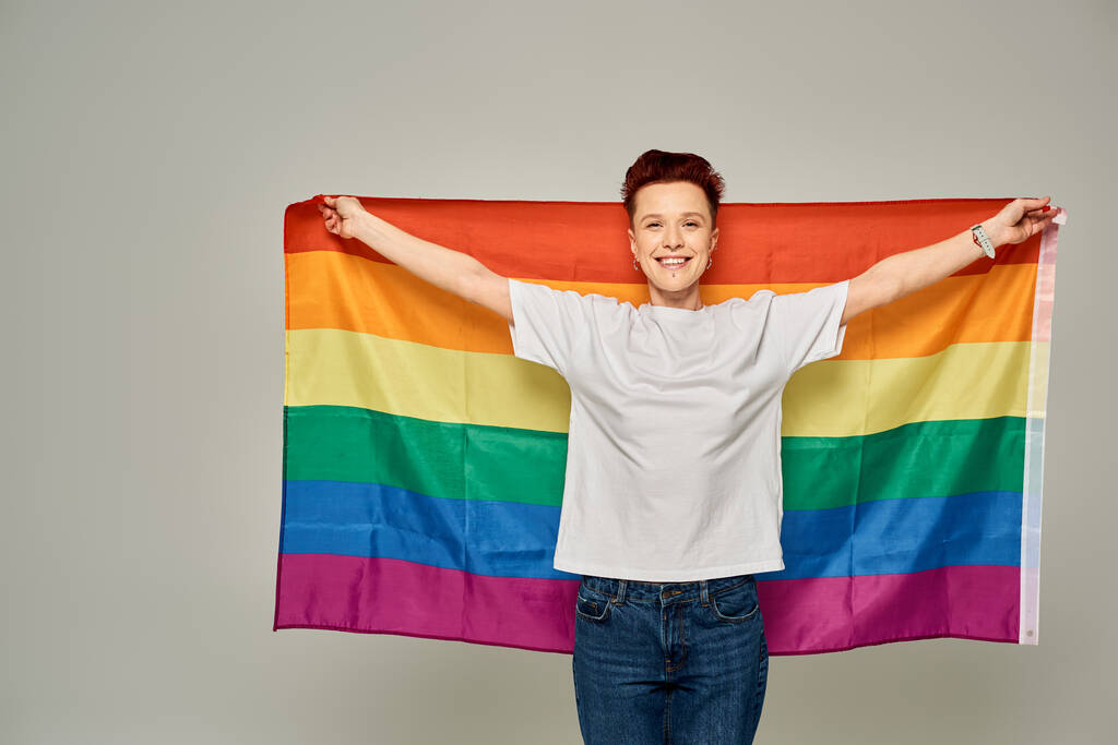 radosna ruda bigender osoba w białej koszulce stojąca z flagą LGBT na szarym tle - Zdjęcie, obraz