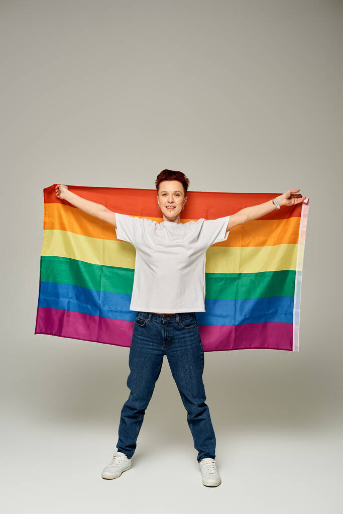 full length of redhead bigender person in white t-shirt and jeans standing with LGBT flag on grey - Photo, Image