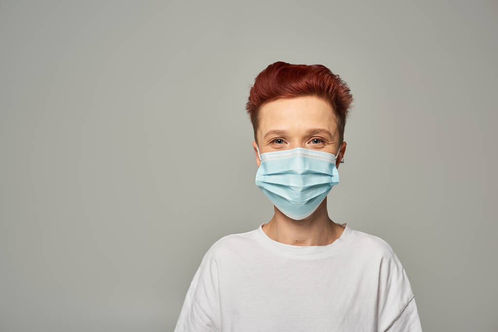 retrato de pelirroja queer persona con mirada feliz con máscara médica y mirando a la cámara en gris - Foto, Imagen