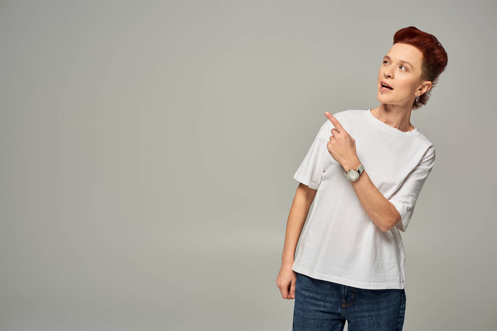 στοχαστική κοκκινομάλλα μη δυαδικό πρόσωπο σε λευκό t-shirt κοιτάζοντας μακριά και δείχνοντας με το δάχτυλο σε γκρι - Φωτογραφία, εικόνα