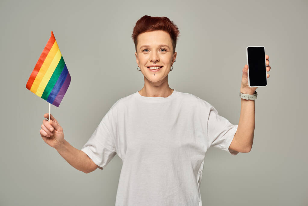 radosna ruda queer osoba trzymająca małą flagę LGBT i smartfon z pustym ekranem na szarym - Zdjęcie, obraz