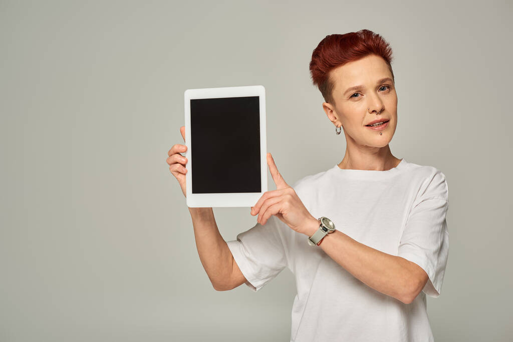 руда небінарна особа в білій футболці, що показує цифровий планшет з порожнім екраном на сірому фоні - Фото, зображення