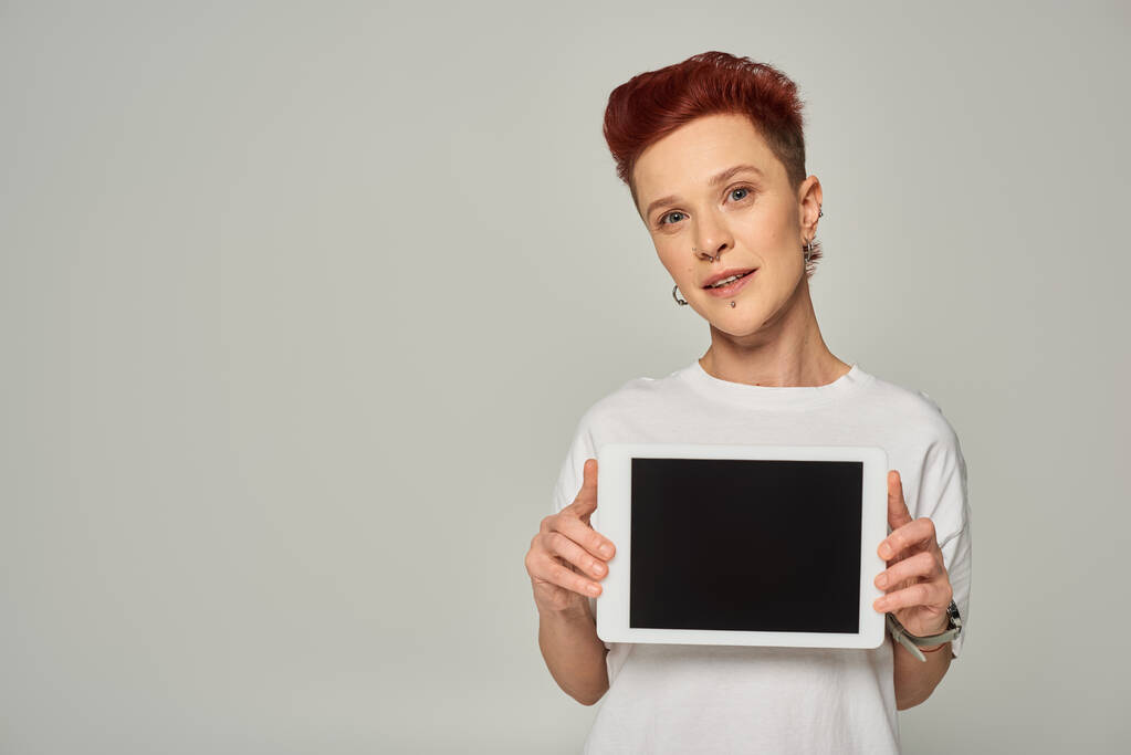 Beyaz tişörtlü, gri arkaplanda boş ekranlı dijital tableti gösteren kızıl saçlı tuhaf kişi. - Fotoğraf, Görsel
