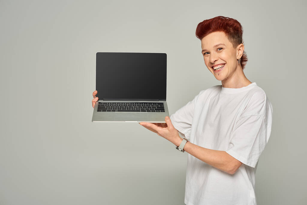 pelirroja queer persona en blanco camiseta mostrando portátil con pantalla en blanco sobre fondo gris - Foto, imagen