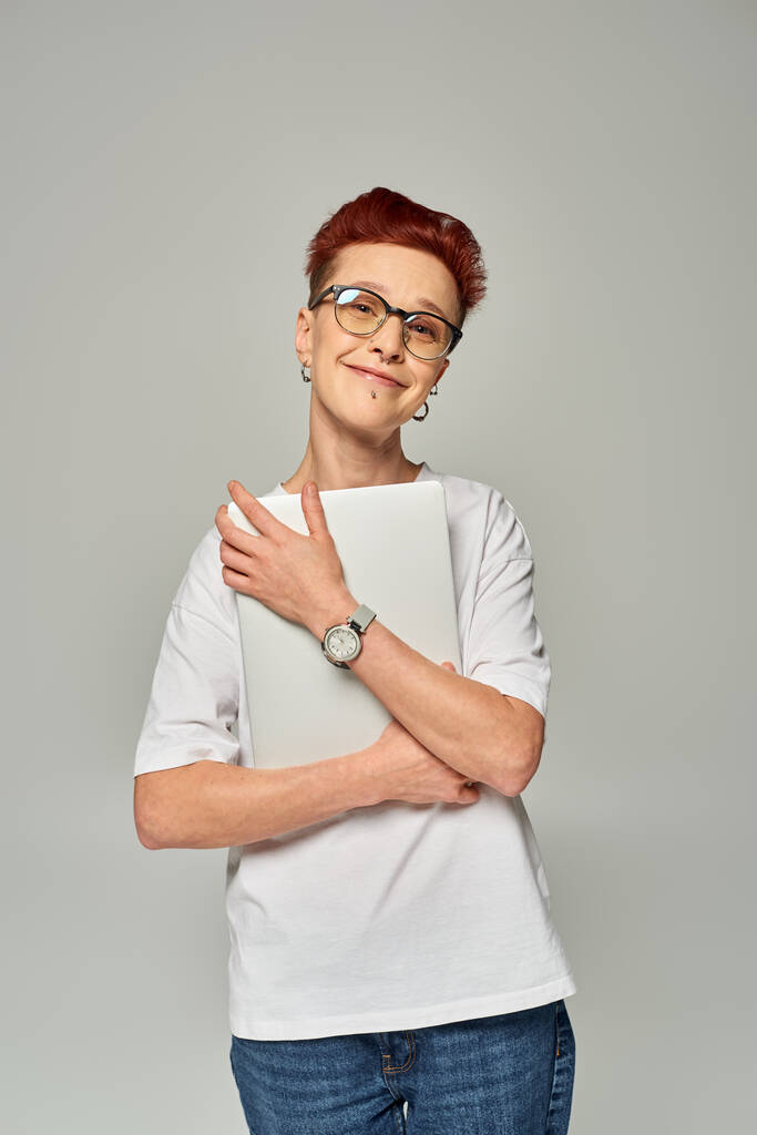 entzückte und glückliche rothaarige queere Freiberuflerin mit Brille und Laptop vor grauem Hintergrund - Foto, Bild