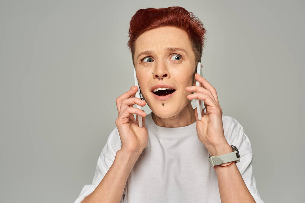 удивленный и удивленный рыжий педик с открытым ртом разговаривающий на смартфонах на сером фоне - Фото, изображение