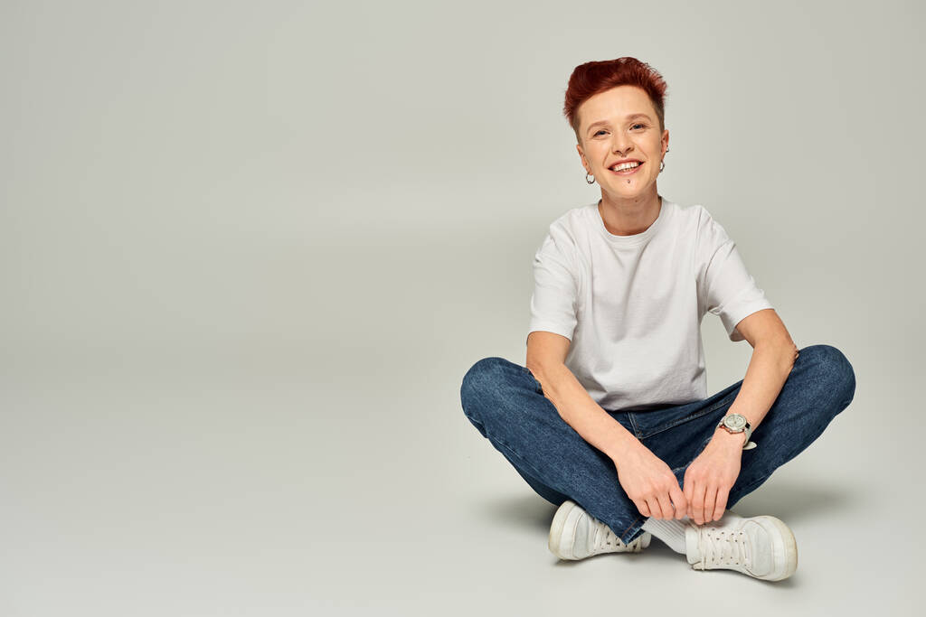 cheerful redhead non-binary person in white t-shirt and jeans sitting and looking at camera on grey - Photo, Image