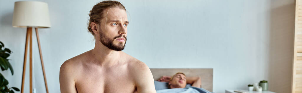 расстроенный бородатый мужчина смотрит в сторону в то время как его любовник спит в спальне, горизонтальное знамя - Фото, изображение