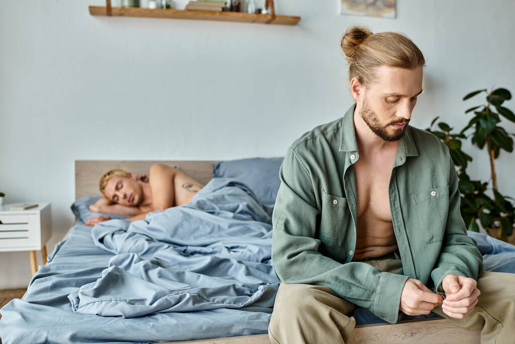 loukkaantunut parrakas homo mies istuu ja pukeutua lähellä poikaystävä makaa makuuhuoneessa aamulla - Valokuva, kuva