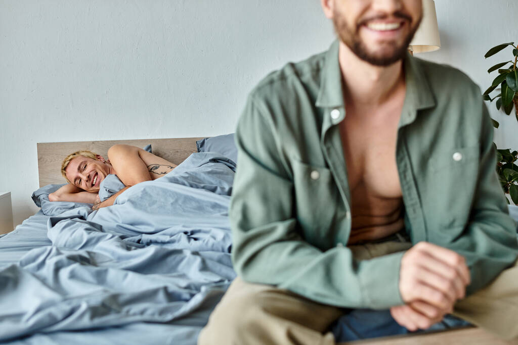 radosny gej człowiek leżący na łóżko i uśmiechając się w pobliżu chłopak siedzi na zamazany pierwszy plan szczęście - Zdjęcie, obraz
