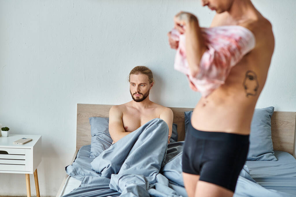 sconvolto barbuto gay uomo seduto sul letto vicino amore partner vestirsi in camera da letto, problemi di relazione - Foto, immagini