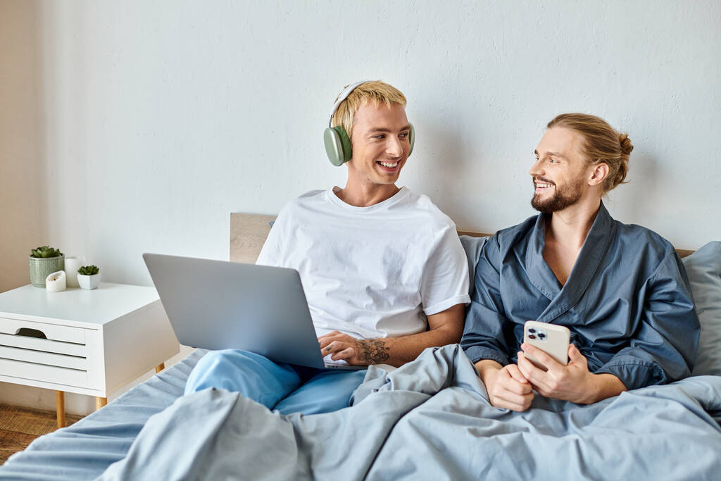 szczęśliwy gej człowiek w słuchawki przeglądanie internet na laptop w pobliżu chłopak z smartphone na łóżko - Zdjęcie, obraz