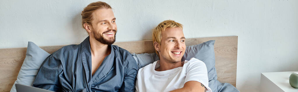 счастливая гей-пара улыбаясь и глядя в сторону, сидя в спальне утром, горизонтальный баннер - Фото, изображение
