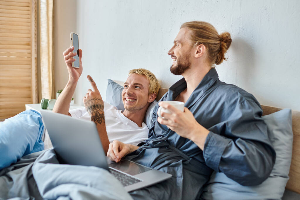 гей чоловік з чашкою кави посміхається біля ноутбука і татуйований хлопець вказує на смартфон на ліжку - Фото, зображення