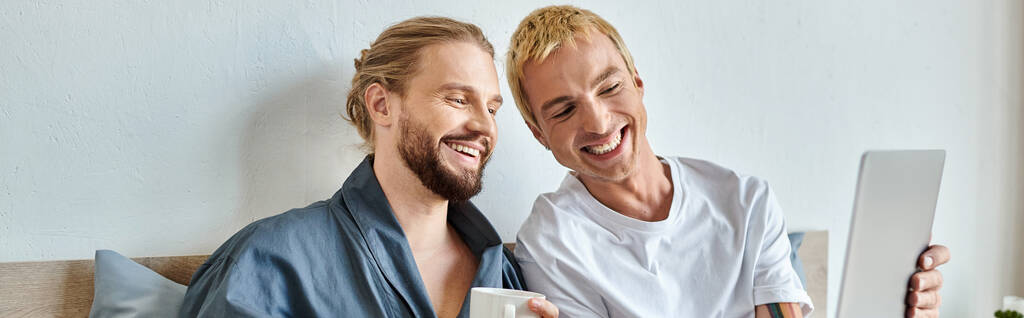 Gülümseyen sakallı sevgiliye dizüstü bilgisayar gösteren neşeli eşcinsel adam yatak odasında kahve içiyor, afiş yapıyor. - Fotoğraf, Görsel