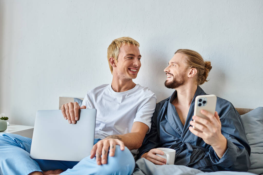 楽しいゲイ男性とコーヒーカップとスマートフォンの笑顔の彼氏とベッドルーム,レジャーでラップトップ - 写真・画像