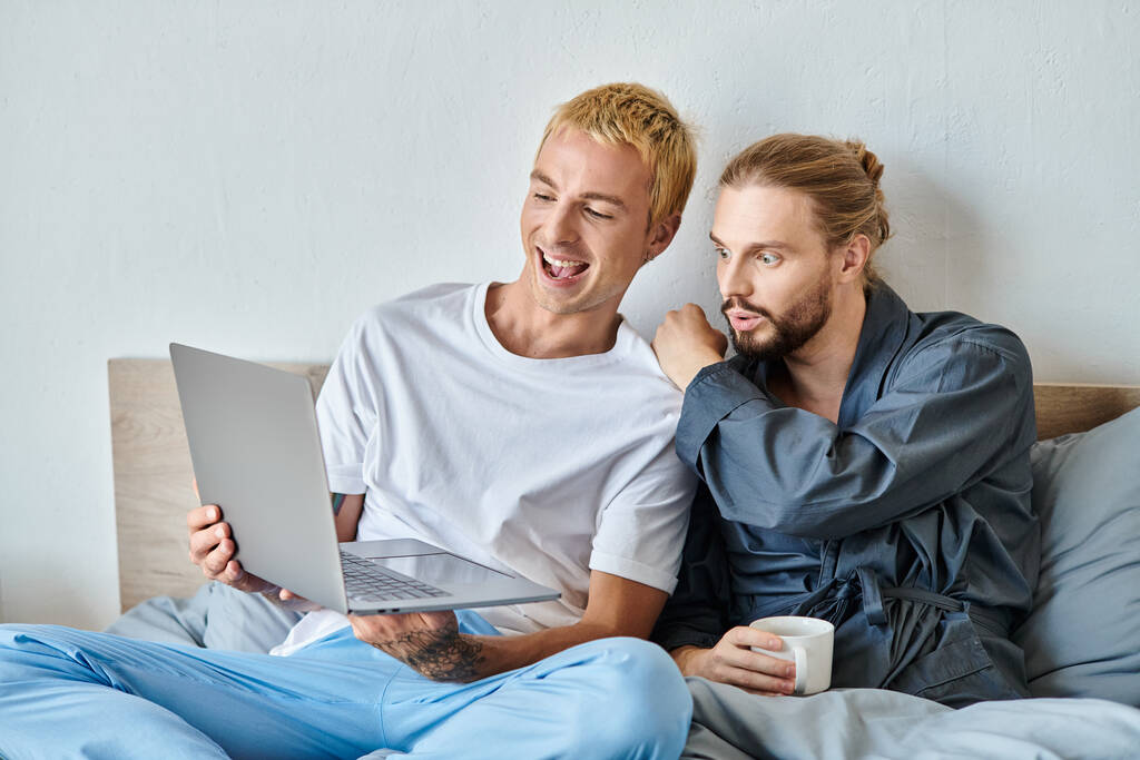 risa gay hombre mostrando laptop a asombrado barbudo hombre sentado con café taza en dormitorio, ocio - Foto, imagen