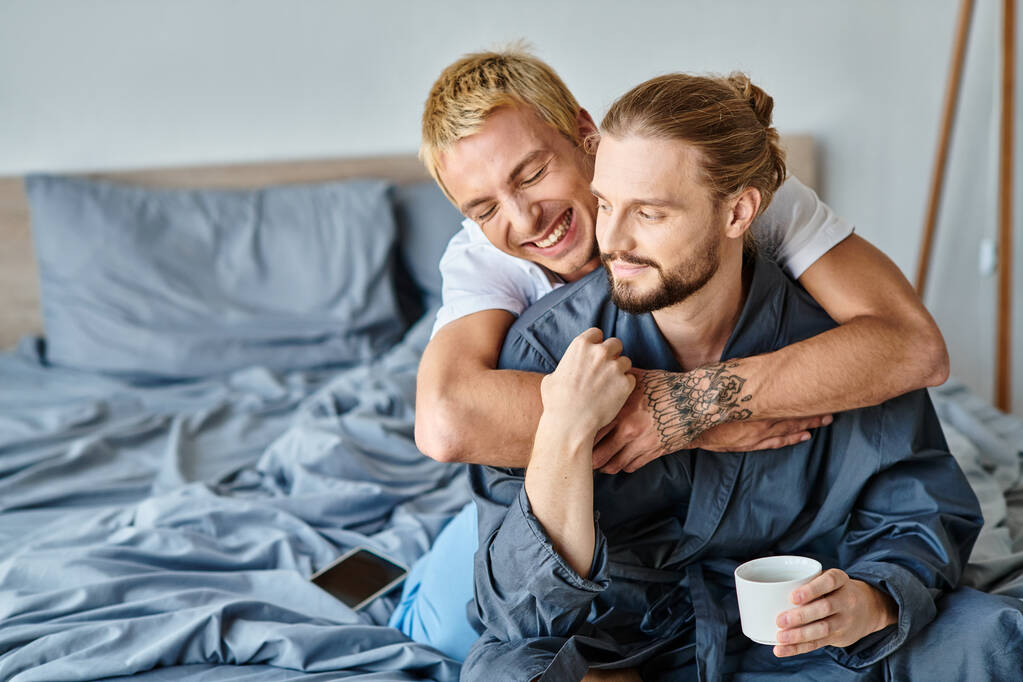 joyful tattooed gay man embracing bearded boyfriend with coffee cup in bedroom, happy scene - Photo, Image