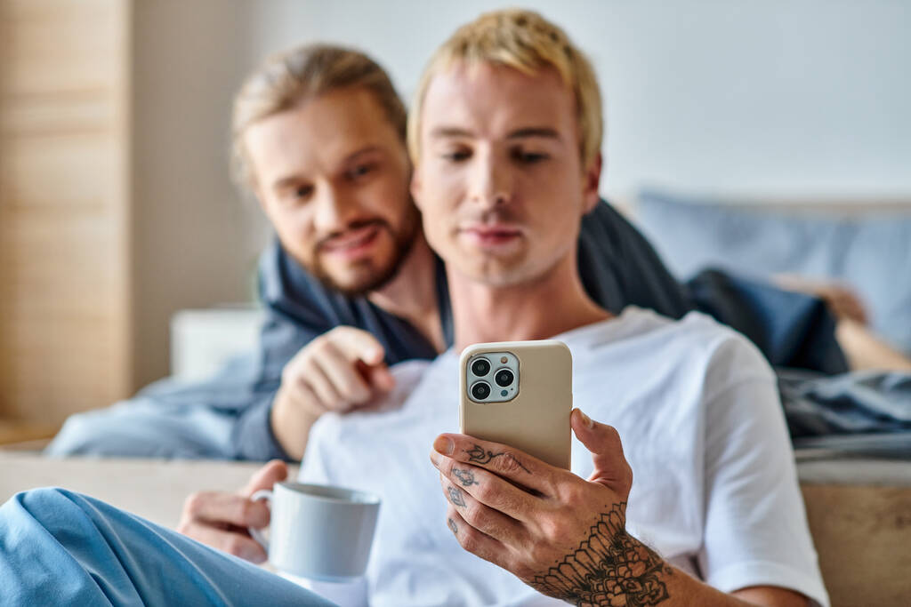 getatoeëerde gay man met koffie kopje browsen internet op mobiele telefoon in de buurt glimlachende vriendje in de slaapkamer - Foto, afbeelding