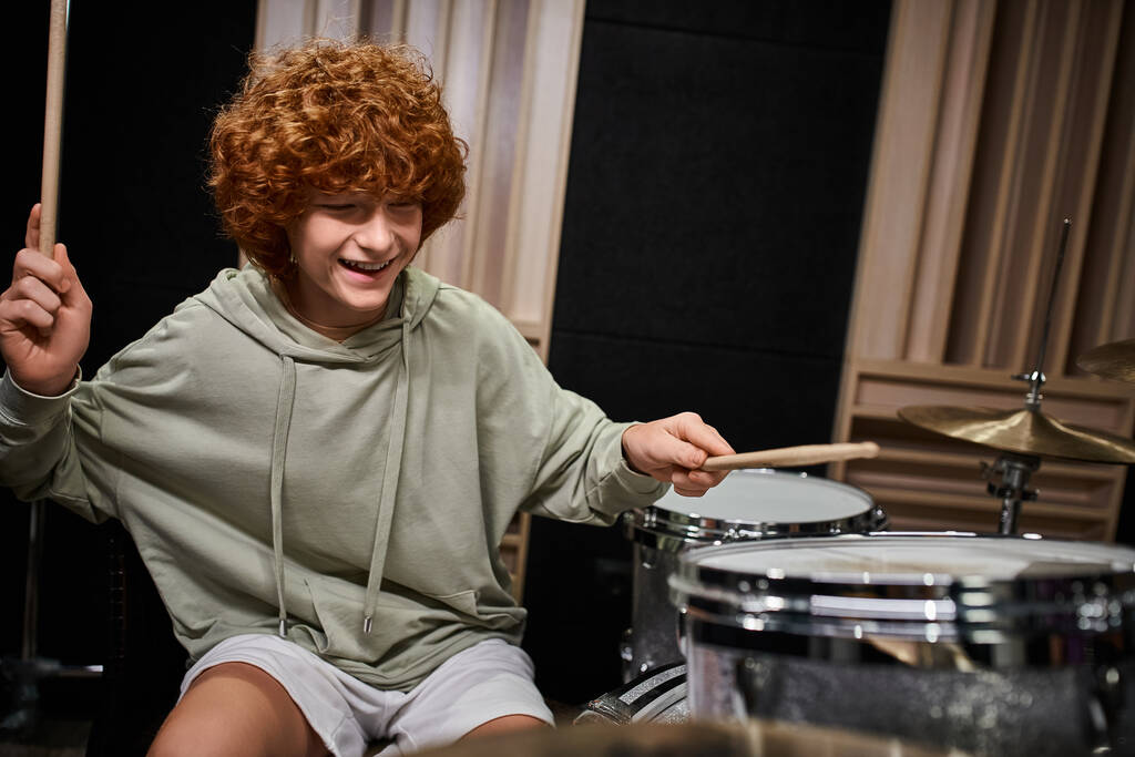 vidám aranyos vörös hajú tinédzser fiú alkalmi öltözékben játszik dob aktívan, míg a stúdióban - Fotó, kép