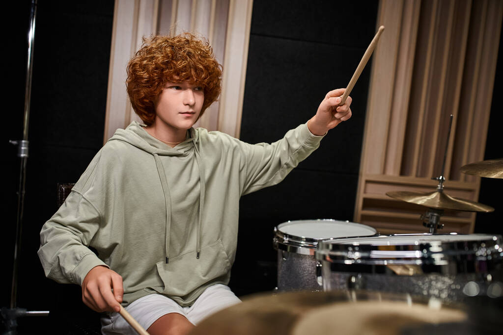 αξιολάτρευτο συμπυκνωμένο έφηβος αγόρι με κόκκινα μαλλιά σε καθημερινή στολή παίζει τύμπανα του, ενώ στο στούντιο - Φωτογραφία, εικόνα