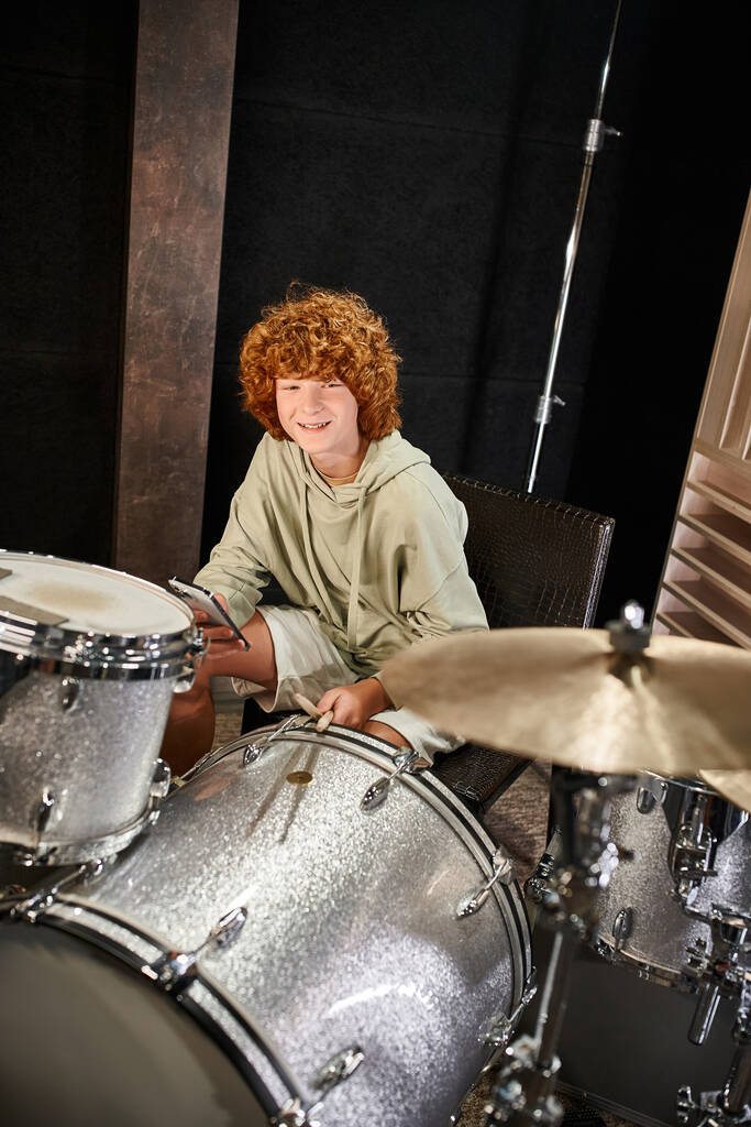 cheerful adorable red haired teenage boy in cozy attire holding mobile phone next to his drums - Photo, Image