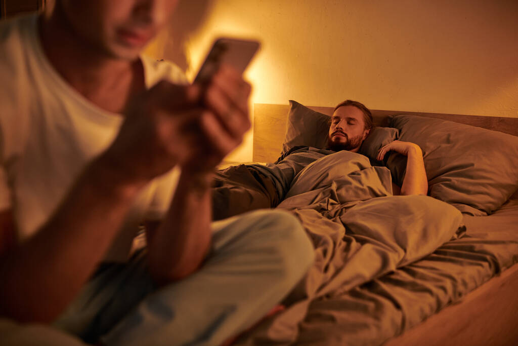 Schwuler Mann schläft nachts, während untreuer Freund auf Smartphone im verschwommenen Vordergrund chattet - Foto, Bild