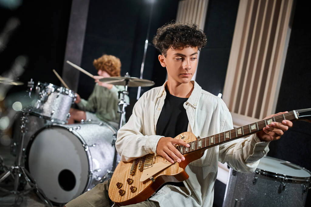 концентрований чарівний хлопчик-підліток в повсякденному вбранні грає на гітарі поруч зі своїм розмитим барабанщиком - Фото, зображення