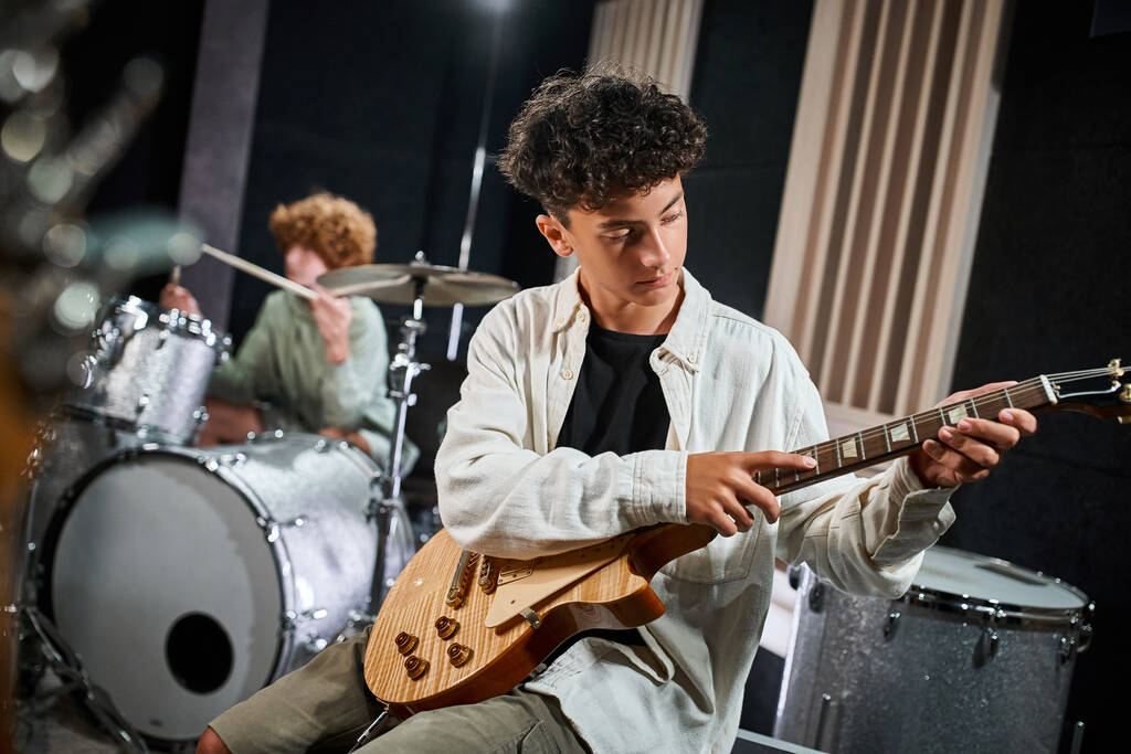 guapo talentoso adolescente tocando la guitarra junto a su borroso baterista, grupo musical - Foto, imagen