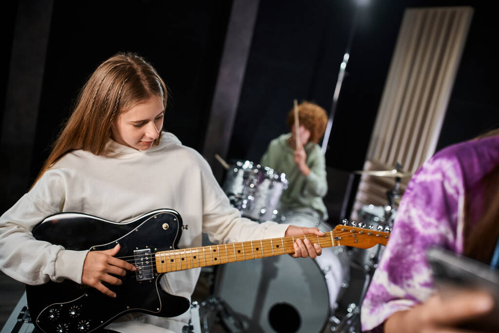adorable rubia adolescente en traje casual tocando la guitarra junto a sus talentosos amigos en el estudio - Foto, Imagen
