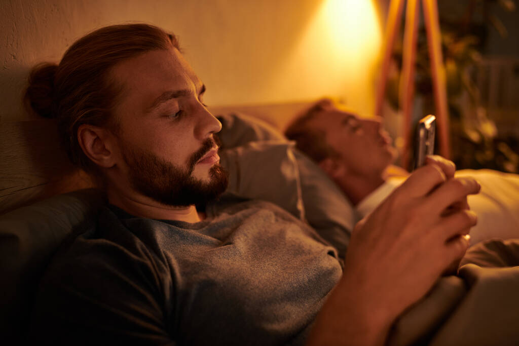 niewierny brodaty gej człowiek wiadomości na telefon komórkowy w pobliżu śpiący chłopak w nocy w sypialnia - Zdjęcie, obraz