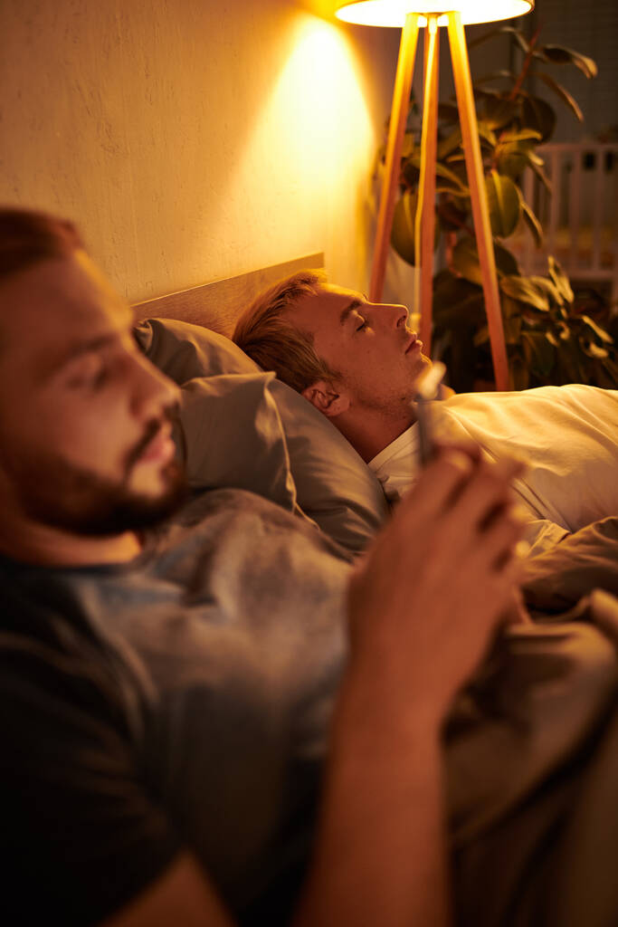 unfaithful gay man browsing date app on mobile phone near sleeping boyfriend at night in bedroom - Photo, Image